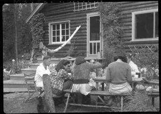 Untitled (group at table outside log cabin)